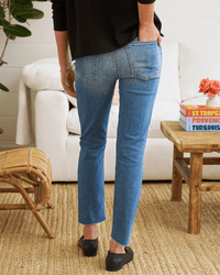 A person in Frank & Eileen's Derry Pant in 2001 Classic Blue Wash and a black top stands with one hand in their pocket, facing away from the camera beside a wooden coffee table and white sofa in a cozy, well-lit room.