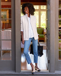 A woman stands in a doorway, smiling and wearing the Frank & Eileen Dublin Knit Blazer in Vintage White over a white shirt, blue jeans, two-tone heels, and holding a black bag. Behind her is a cozy, sunlit room with large windows and furniture.