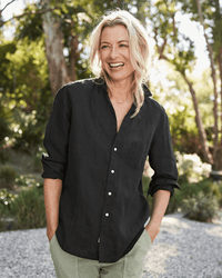 A smiling blonde woman wears the Frank & Eileen Eileen Relaxed Button Up in Black Performance Linen with light pants, standing outdoors with hands in pockets amid sunlit green trees and a natural backdrop.
