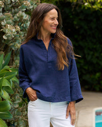 A woman with long brown hair, wearing a Frank & Eileen Gabi Popover Shirt in Navy Linen and white pants, stands outdoors near green plants, smiling and looking to the side.