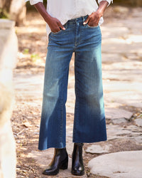 A person stands outdoors on a stone path, hands in pockets, wearing Frank & Eileen Galway Gaucho in 1999 Blue Wash jeans, black ankle boots, and a white shirt. The background features blurred natural elements and soft sunlight.