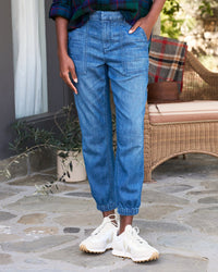 A person stands on a stone patio wearing Frank & Eileen’s Jameson Utility Jogger in 1984 Washed Blue, white sneakers, and a dark plaid shirt. A wicker bench with a plaid blanket and a potted plant are behind them. Their head is not visible.