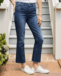 A person stands outdoors on a brick pathway in front of stairs, wearing Frank & Eileen's Killian Crop Flare Jean in 2003 Medium Wash, a white shirt, white sneakers, and a bracelet, with one hand resting on their thigh.
