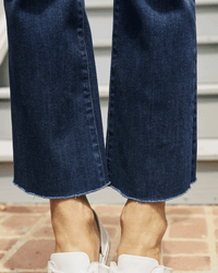 A close-up of a person wearing Frank & Eileen's Killian Crop Flare Jean in 2003 Medium Wash with raw, frayed hems and white shoes, standing on a brick surface in front of steps.