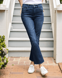 A person stands on a brick walkway before stairs, wearing Frank & Eileen’s Killian Crop Flare Jean in 2003 Medium Wash, a white shirt, and white sneakers. Their hands are in their pockets and their head is not visible.