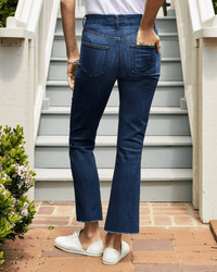 A person stands outside on a brick path, facing away, wearing Frank & Eileen’s Killian Crop Flare Jean in 2003 Medium Wash with frayed hems, white sneakers, and a white top; stairs and greenery are in the background.