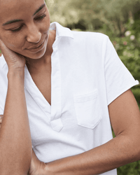 Someone stands outdoors against a blurred green background, wearing the Frank & Eileen Lauren S/S Polo Jersey Dress in White with a breast pocket, looking down and touching their neck.