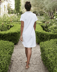 A woman in a Frank & Eileen Lauren S/S Polo Jersey Dress in White walks barefoot along a gravel path through a vibrant garden with trimmed hedges and trees, photographed from behind.