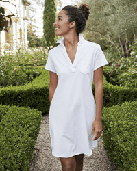 A woman in the Lauren S/S Polo Jersey Dress in White by Frank & Eileen stands on a gravel path in a lush garden, smiling amid green hedges and trees, highlighting the Cotton Heritage Jersey fabric.