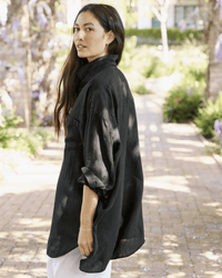 A woman with long dark hair stands outdoors on a sunlit brick path, wearing Frank & Eileen's Mackenzie in Black Performance Linen. Looking over her shoulder, the lush green background highlights her effortless style.