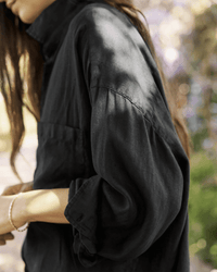 A person with long brown hair wears the Frank & Eileen Mackenzie in Black Performance Linen outdoors in dappled sunlight. Their face isn’t visible, and they have a thin bracelet on their wrist.