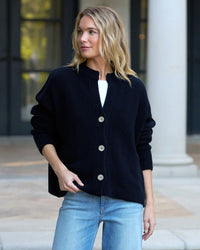A woman with blonde hair stands outdoors before columned architecture, wearing a Frank & Eileen Montecito Cardigan in Marine over a white top and light blue jeans.