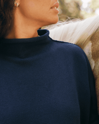 A woman, with only her lower face, neck, and upper torso visible, stands next to a horse while wearing the Frank & Eileen Monterey Sweater in Navy. The horse’s neck and mane appear in the background.