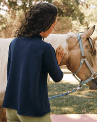 Wearing the Frank & Eileen Monterey Sweater in Navy, a person with curly hair gently pets a light brown horse with a white mane and blue halter outdoors on a sunny day.