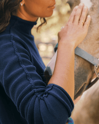 Wearing a Frank & Eileen Monterey Sweater in Navy, a person gently touches a horse’s face, creating a calm, intimate scene. The person's face is partially visible and the horse wears a halter.