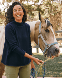 A smiling woman with curly hair wears the Frank & Eileen Monterey Sweater in Navy as she stands outdoors beside a light brown horse with a blue halter, holding its reins and grass for it to eat, with trees and a fence in the background.