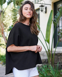 A woman with long brown hair stands confidently outdoors near greenery and a white building, wearing Frank & Eileen's Olive Capelet in Black and white shorts.