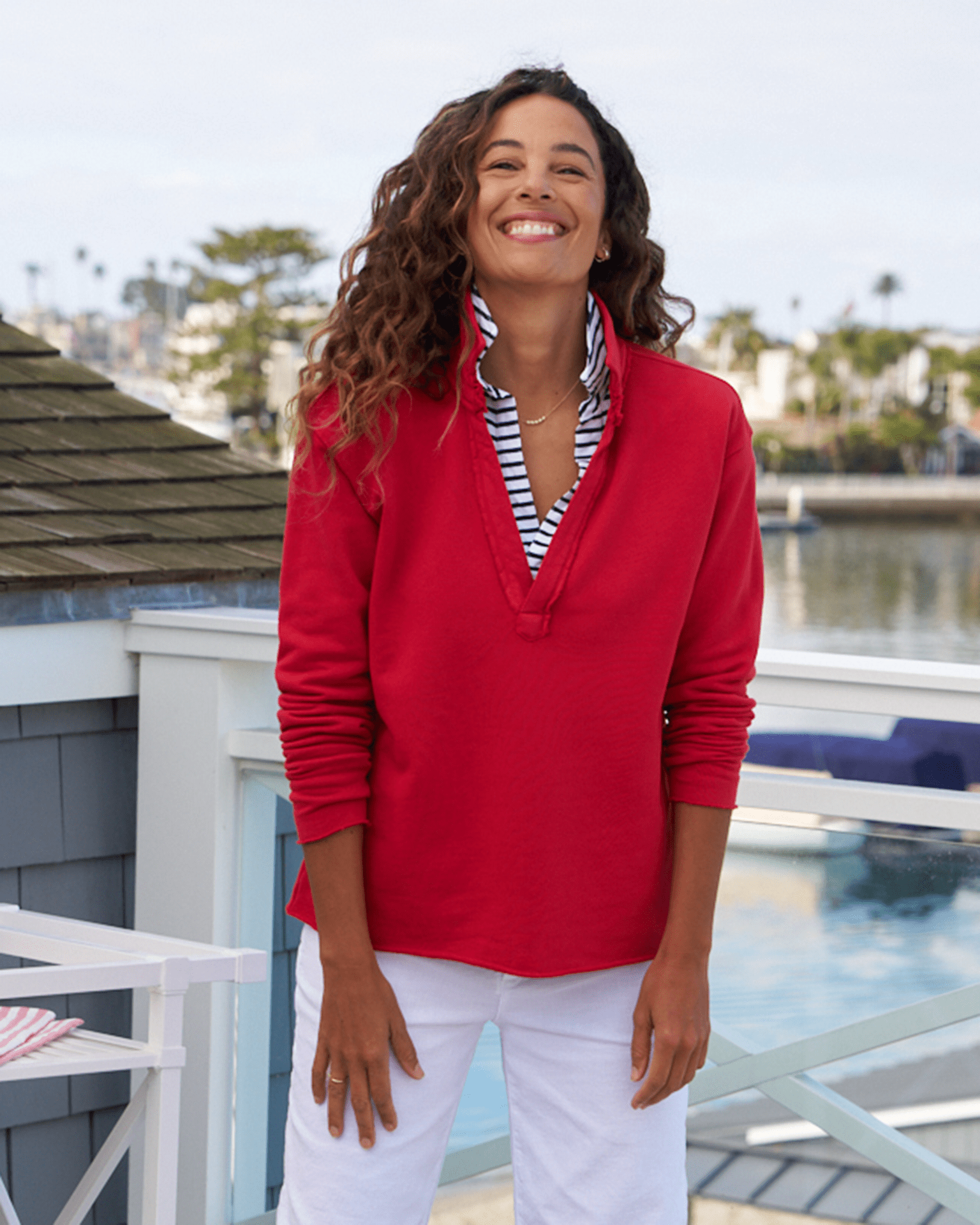 A person in a Double Decker Red French Terry Patrick Popover Henley and white pants stands smiling on a deck, with a body of water and buildings in the background, embodying the effortlessly chic style of Frank & Eileen.