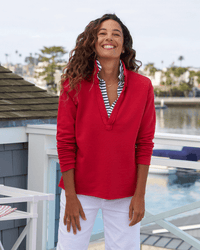 A person in a Double Decker Red French Terry Patrick Popover Henley and white pants stands smiling on a deck, with a body of water and buildings in the background, embodying the effortlessly chic style of Frank & Eileen.