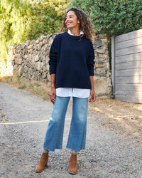 A woman smiles outdoors on a dirt path, wearing the Frank & Eileen Montecito Sweater in Marine over a white shirt, with cropped blue jeans and brown ankle boots. Behind her are a stone wall, lush greenery, and a wooden gate.