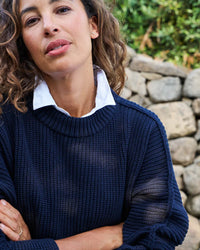 A woman with curly brown hair wears the Frank & Eileen Montecito Sweater in Marine over a white collared shirt, standing before a stone wall and greenery, and looking to the side with her arms crossed.