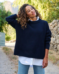 A woman with curly hair stands on a dirt path in front of greenery and a stone wall, wearing the Frank & Eileen Montecito Sweater in Marine over a white shirt and light blue jeans, looking to the side with one hand touching her hair.