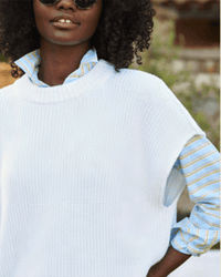 A person with curly hair wears the Frank & Eileen Montecito Vest in White over a blue and yellow striped shirt outdoors, showing only their upper body and part of their face.