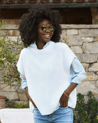A smiling woman with curly hair wears sunglasses and the Frank & Eileen Montecito Vest in White over a striped shirt and jeans, standing outdoors in front of a stone wall and green plants.