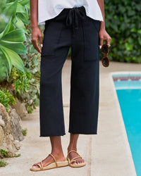 By a pool, a person wears Frank & Eileen’s Penny Patch Pocket Sweatpant in British Royal Navy with a white top and tan sandals, holding sunglasses. Lush green plants fill the background.