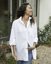 A woman with curly hair, wearing the Frank & Eileen Shirley Oversized Button Up in White Everyday Poplin and blue jeans, stands outdoors on a gravel path, smiling to the side with plants and a building in the background.
