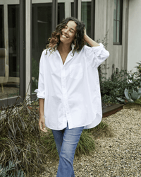 A woman with curly hair smiles outside a modern building, wearing the Frank & Eileen Shirley Oversized Button Up in White Everyday Poplin and blue jeans, with one hand touching her hair.