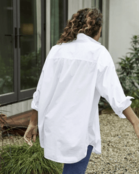 A person with wavy hair, wearing the Frank & Eileen Shirley Oversized Button Up in White Everyday Poplin and blue jeans, walks away on a gravel path near a modern glass door and greenery.