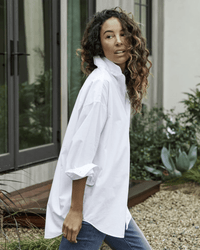 A woman with curly hair wears the Shirley Oversized Button Up in White Everyday Poplin by Frank & Eileen, styled with blue jeans. She stands outdoors near a modern building enhanced by plants and gravel accents.