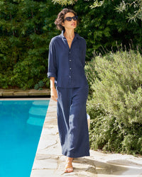 A woman in sunglasses wears the Frank & Eileen Tuscany Linen Set in Navy, featuring a button down shirt and matching wide leg pants, as she walks by a swimming pool with lush greenery in the background.