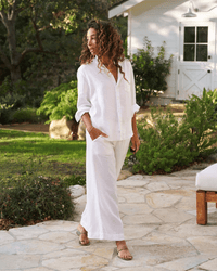 A woman with curly hair stands barefoot on a stone patio in Frank & Eileen’s Tuscany Linen Set in White, featuring a relaxed button down shirt and wide leg pants, surrounded by lush greenery near a white cottage.