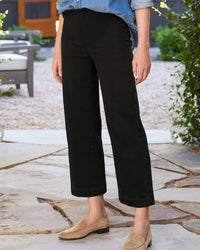 Standing outdoors on a stone patio with greenery and patio furniture, a person wears Frank & Eileen’s Westport Wide Leg Illusion in Black—cropped denim with a pull-on waistband—paired with a light blue rolled-sleeve shirt and beige loafers.
