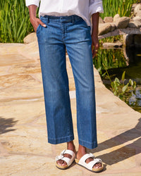 A person stands outdoors on stone pavement near water, wearing Frank & Eileen’s Westport Wide Leg in 1984 Washed Blue Wash jeans, a white long-sleeve shirt, and white bow sandals. Their hands are in their pockets, and their face is not visible.
