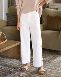 A person wears a light pink sweater, Frank & Eileen’s Wexford Wide-Leg Linen Pant in White Linen, and beige slide sandals while standing indoors on a patterned rug near a wooden coffee table and an olive green sofa.