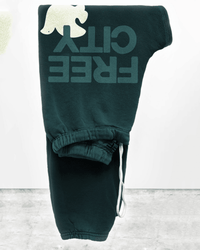 The FC Large Sweatpants in Greenshrooms by Free City are dark green French terry sweatpants with "FREE CITY" and a white dove graphic on the thigh, featuring an elastic waistband and white drawstrings, displayed on a light background.