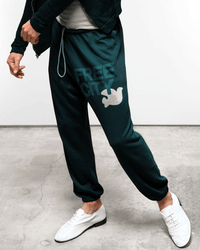 A person stands indoors wearing Free City FC Large Sweatpants in Greenshrooms with a dove graphic on the left thigh, paired with white shoes and a dark jacket. The setting features a concrete floor and plain white background—ideal for stylish loungewear.