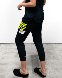 A person stands on a concrete floor against a white wall, wearing Free City Large 3/4 Sweatpants in Squidink Lemons, which are black with yellow "SWEETY" lettering and a white bunny graphic, along with a black shirt and black slippers.
