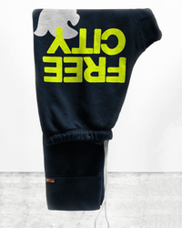 The Free City Large 3/4 Sweatpants in Squidink Lemons feature bold yellow FREECITY text and a white dove graphic. Handmade in LA from 100% cotton, these dark sweatpants are folded on a gray surface for a premium comfort look.