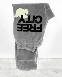 A pair of Free City Large 3/4 Sweats in Heather Cream, handmade in LA with an upside-down yellow dove logo, folded on a gray marble surface against a white background.