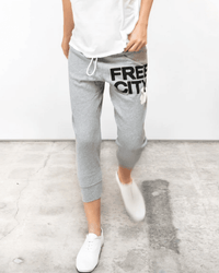 A person wears Free City Large 3/4 Sweats in Heather Cream, a white t-shirt, and white sneakers, standing on a concrete floor with a plain white background.