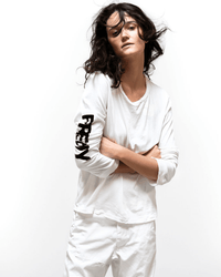 A person with wavy dark hair wears the Free City LNLSun Longsleeve T in Creamy Yum, featuring FREEDOM printed in black on the sleeve, paired with white pants and standing against a plain white background with arms crossed.