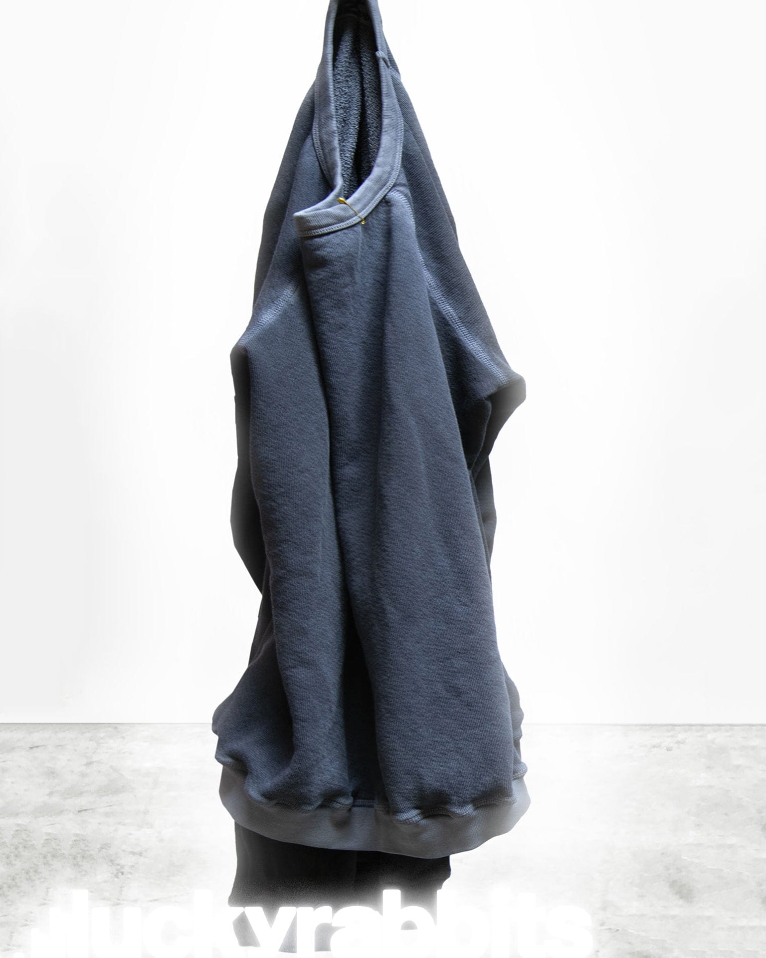 The Free City Lucky Rabbits Sweatshirt in Moonshroom Blue, a dark blue French terry hoodie, hangs on a hook against a plain white background with "lucky rabbits" faintly visible at the bottom.