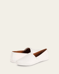 A pair of FRYE Melanie Slip On sneakers in white, featuring vintage leather details and brown lining. One shoe stands upright, the other lies on its side, both set against a plain off-white background.