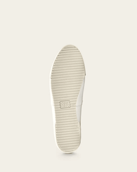 The image displays the bottom view of the Melanie Slip On in White by FRYE, featuring a ridged sole with the FRYE logo embossed at the center. The background is solid light beige.