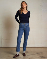 A woman stands indoors by a light wall, wearing the Georgina Cashmere V Neck Sweater in Navy from perfectwhitetee, blue high-waisted jeans, gray socks, and brown loafers. Her hands are in her pockets as she gazes slightly upward.