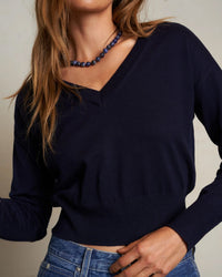 A woman wears the perfectwhitetee Georgina Cashmere V Neck Sweater in Navy with blue jeans and a beaded necklace, one hand on her hip. Her face is partially out of frame.